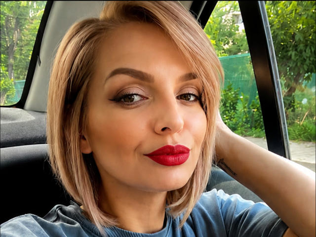 DouceAmelia has blonde bob, red lipstick, and smoky eye makeup, smiling with hand behind head in car, wearing blue shirt, showing cute ass in background.