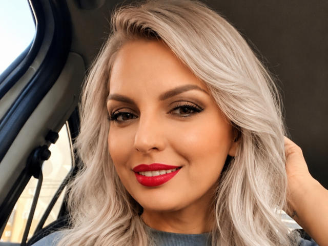 DouceAmelia with silver hair, red lipstick, and black eyeliner, smiling in a car, hand in hair, looking cute and sexy, with a confident and sexy vibe.