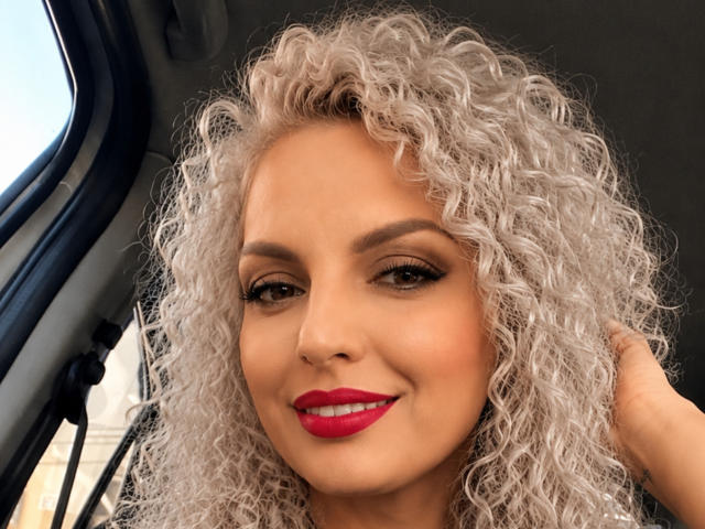 DouceAmelia with curly white hair, tan skin, red lipstick, and brown eyes, smiling in a car, hand touching her head. Her tits aren't visible in the photo.