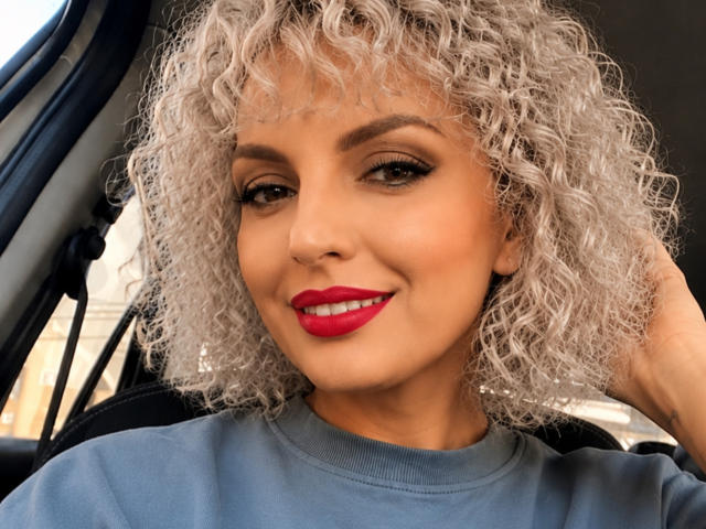 DouceAmelia with curly silver hair, red lipstick, and gray sweater, smiling in a car, looking sexy with her hand in her hair. Her tits are not visible.