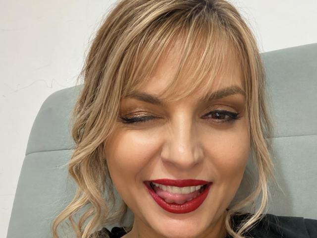 DouceAmelia's face, winking with right eye, has blonde hair, red lipstick, and black eyeliner. Her tongue is slightly out, showing her teeth. She's sitting on a grey chair.