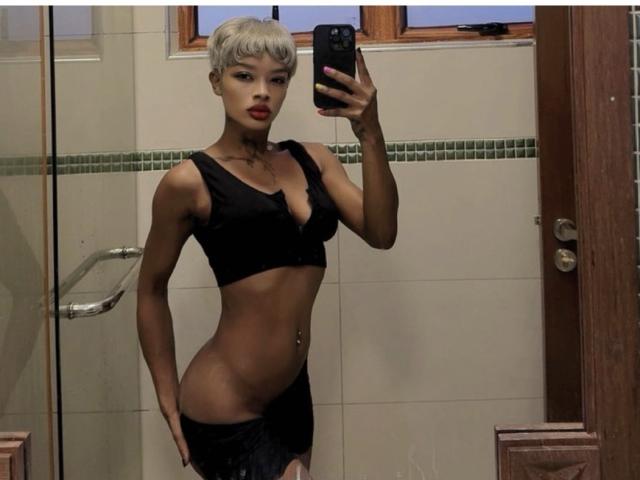 Halinah, con cabello rubio platino corto, vestida con un sostén negro y pantalones negros ajustados, mostrando su abdomen plano y su piercing en el ombligo, tomando una selfie frente al espejo con su teléfono..