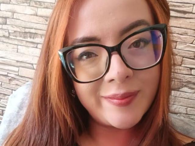 AntonellaRodika ha i capelli rossi, gli occhiali neri e un leggero sorriso con labbra rosa. Il suo sedere non è visibile, si vede solo il suo viso. Sta guardando la fotocamera..