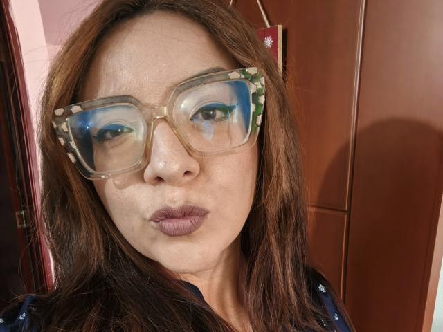 Antonella Rodika, con i capelli castani, indossa occhiali trasparenti con lenti riflettenti che mostrano il suo viso e rossetto viola, con le labbra strette. Indossa una camicia blu scuro..