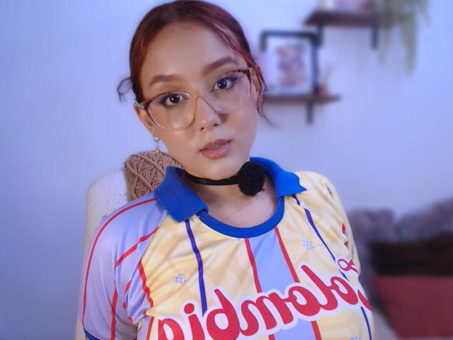 Sophia Browm, com cabelo ruivo, óculos e um fone de ouvido preto, veste uma camisa amarela e azul com a inscrição "idmmor" e tem uma expressão séria. Seus seios são pequenos e ela está olhando para a câmera..