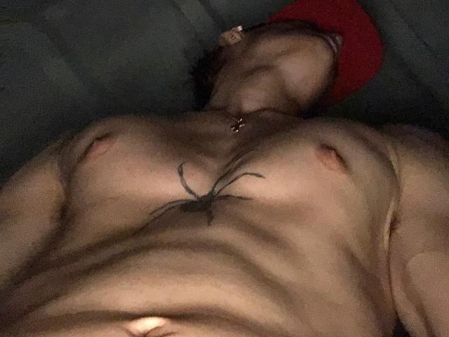 SebasSexyMan está acostado, sin camisa, con un tatuaje entre sus pezones, un sombrero rojo en la cabeza y el ombligo visible. Sus pezones están ligeramente erectos..