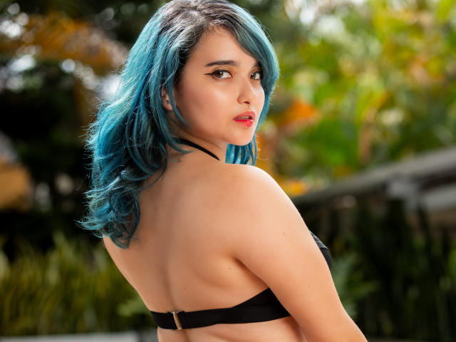 Naommy Carter, com cabelo azul, olhando por cima do ombro, vestindo um sutiã preto, mostrando suas costas e uma pequena nádega, em pé ao ar livre, com luz natural, com um leve sorriso..
