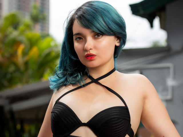 NaommyCarter with blue hair, wearing a black strapless bra, showing her tits, has red lipstick, and a black choker, standing outdoors, looking at the camera.