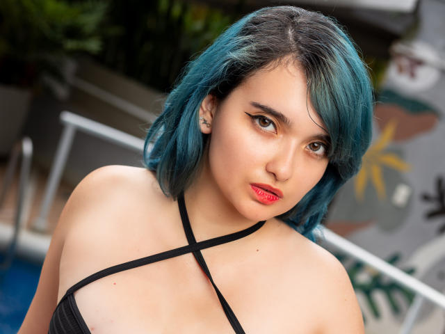 Naommy Carter mit türkisfarbenem Haar, rotem Lippenstift, einem schwarzen Oberteil mit Kreuzungsdetails, das ihre Brüste zeigt, blickt ernst in die Kamera, am Pool, tagsüber..
