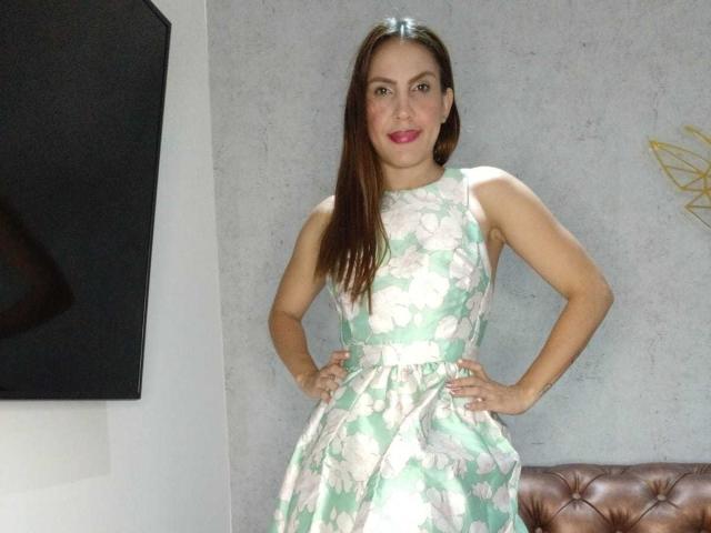 Susan Thomass, con le mani sui fianchi, indossa un abito verde con fiori, ha lunghi capelli castani, rossetto rosa e un sorriso sicuro, ed è in piedi accanto a un divano marrone con decorazioni..
