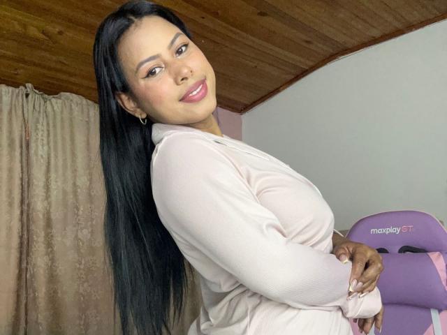 MichHt, com cabelo preto longo, batom rosa e uma blusa rosa de manga comprida, sorrindo para a câmera, mostrando o bumbum enquanto está em um quarto com teto de madeira..