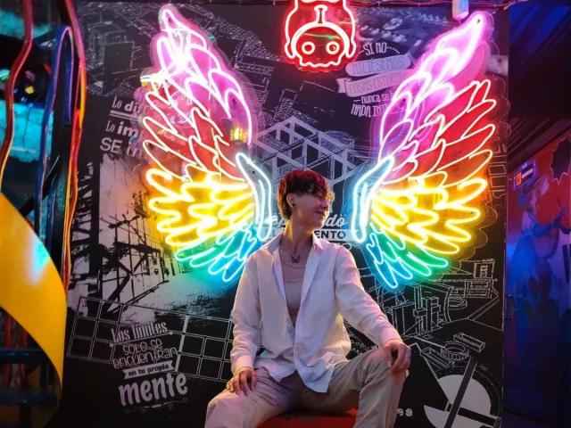 LissyLuxe, sitting with legs apart, wearing a white suit, neon rainbow wings behind, dark background with graffiti, colorful lights, and a red neon head symbol above.