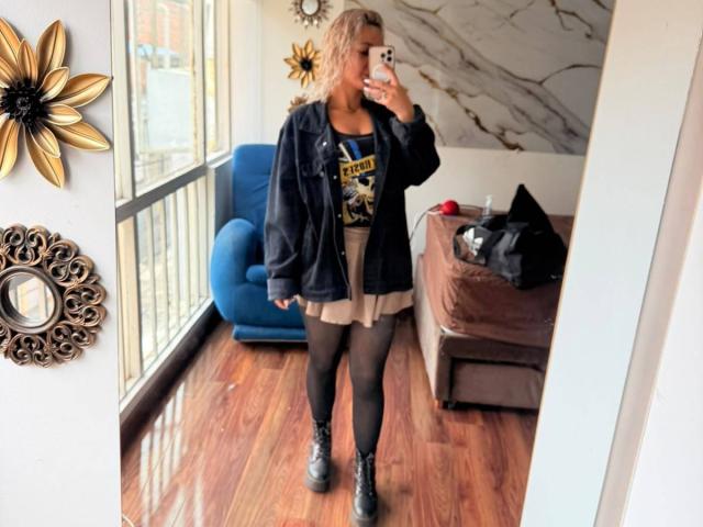 Elena Saenz, avec ses cheveux blonds, sa veste noire, son t-shirt à motif, sa jupe beige, ses collants noirs et ses bottes noires, prend un selfie devant un miroir, montrant légèrement ses fesses et ses seins visibles sous la jupe..
