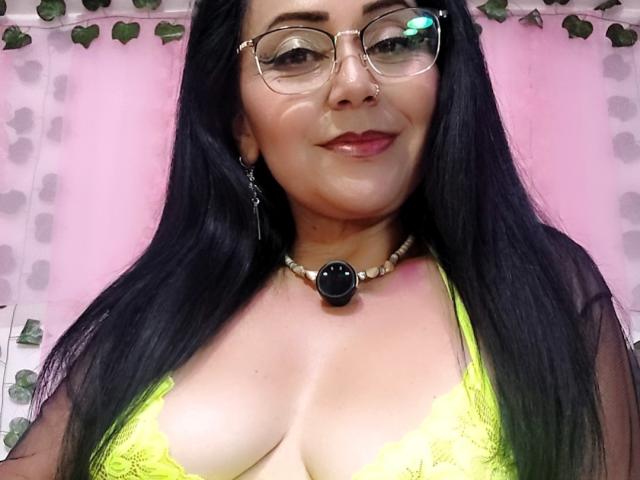 Zaria69, con lunghi capelli neri, occhiali e piercing al naso, indossa un reggiseno di pizzo giallo, una collana con una pietra nera e una maglia trasparente, sorridendo alla fotocamera e mostrando il seno..