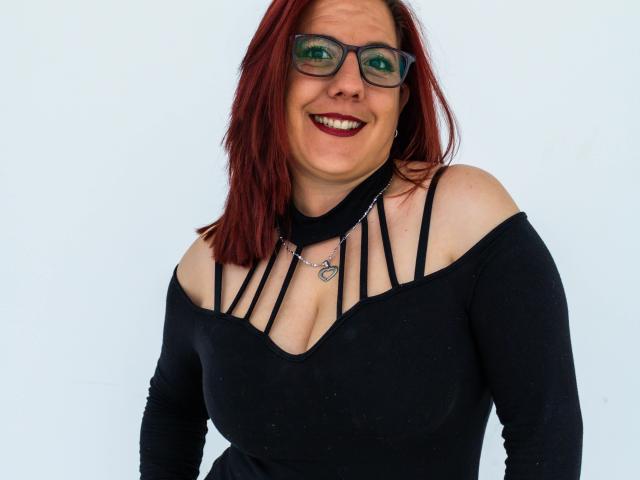 JessyJoness con capelli rossi, occhiali, top nero con spalline scoperte che mostra il décolleté, sorridente, rossetto scuro, collana, sfondo bianco. Il seno è visibile attraverso il top con spalline..