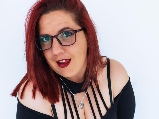 JessyJoness tiene el pelo rojo, gafas y un lápiz labial rojo oscuro. Lleva un top negro con tirantes que deja ver parte del escote, y un collar de plata, todo sobre un fondo blanco..