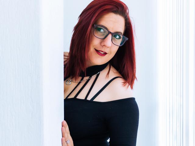 JessyJoness with red hair, glasses, black off-shoulder top, strappy neckline, ring, and red lipstick, peeking from behind a white pillar. Her tits are visible. She's smiling.