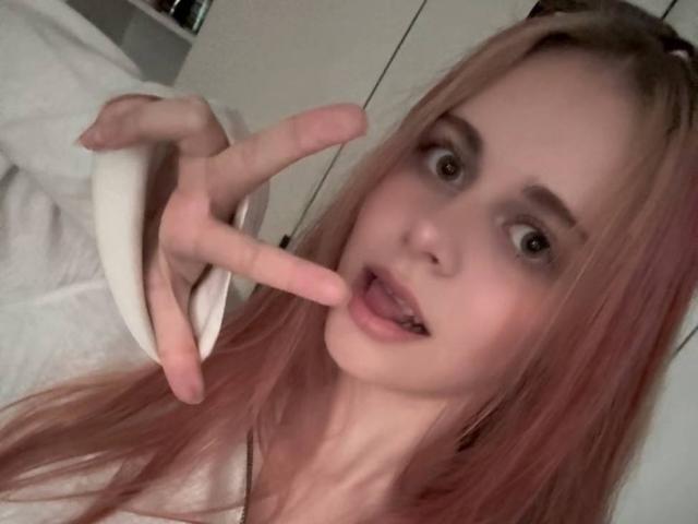 KarenaRomani, con cabello rosa, piel clara y ojos marrones, hace el gesto de la paz con su mano derecha, con la lengua ligeramente afuera, y lleva puesto un top blanco..