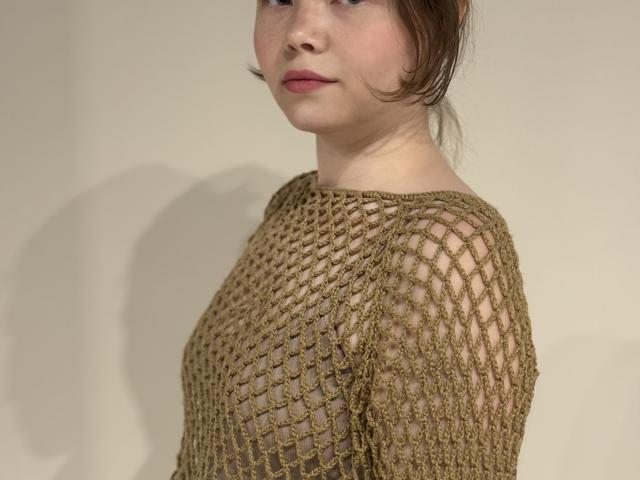DaynaTays, com pele clara, cabelo castanho preso em um coque desarrumado, vestindo uma blusa de malha marrom transparente que mostra seus pequenos seios e mamilos. Batom rosa claro, expressão neutra..