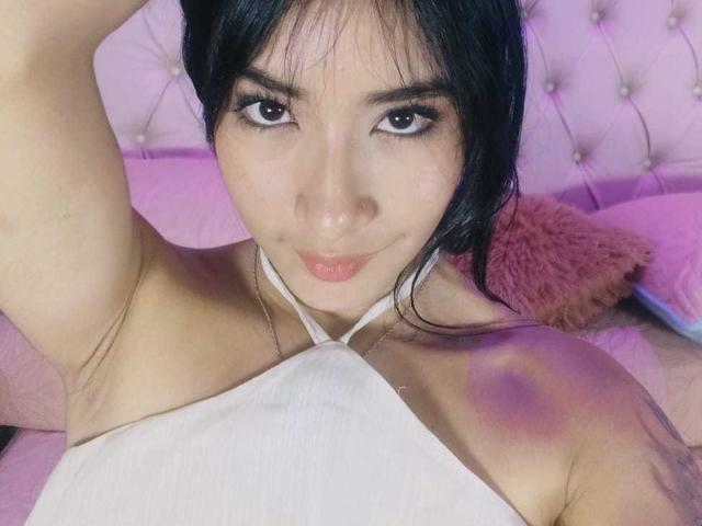 KatheLeclecr with dark hair, brown eyes, and pink lipstick, wearing a white halter top, has a small bruise on her shoulder, lying on a pink bed.