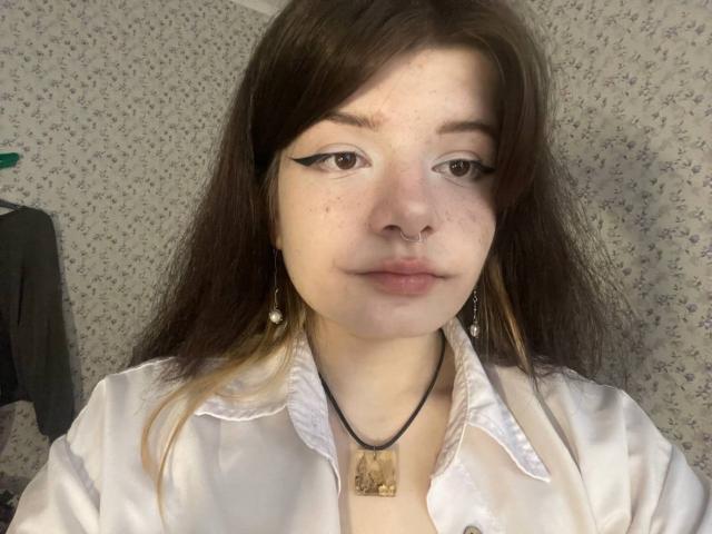 Marla Rotan, met bruine ogen, zwarte eyeliner, een neusring, parel oorbellen, een wit shirt, een zwarte ketting en bruin haar met blonde punten, kijkt naar de camera met een neutrale uitdrukking..