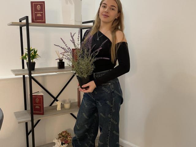Imelda Prattis è in piedi, con un mazzo di lavanda in mano, indossa una maglietta nera con una spalla scoperta e pantaloni di jeans a vita alta, ha lunghi capelli biondi ed è accanto a una mensola con piante e libri..