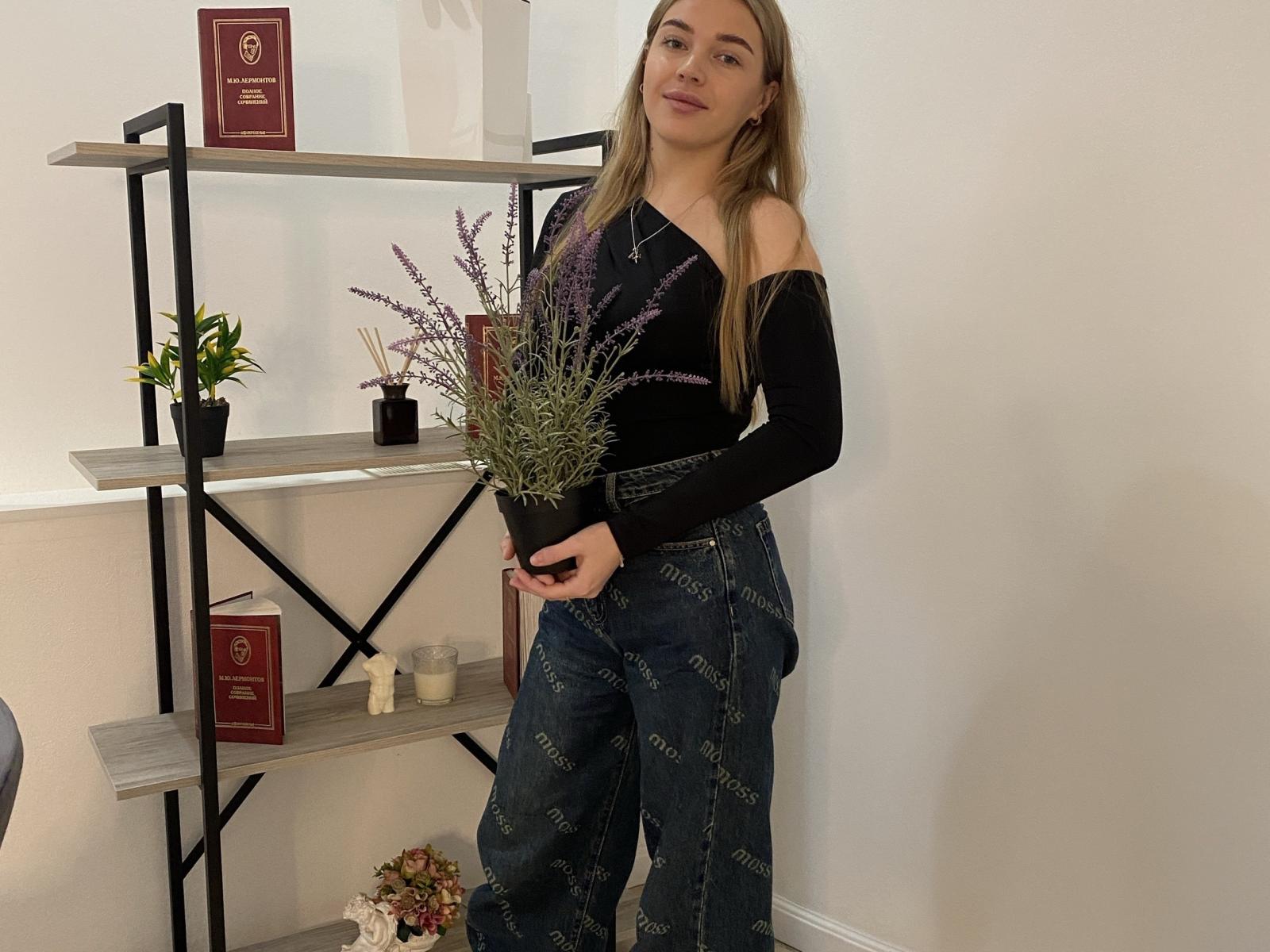 ImeldaPrattis stands holding lavender, wearing a black off-shoulder top and high-waisted denim pants, with long blonde hair, next to a shelf with plants and books.