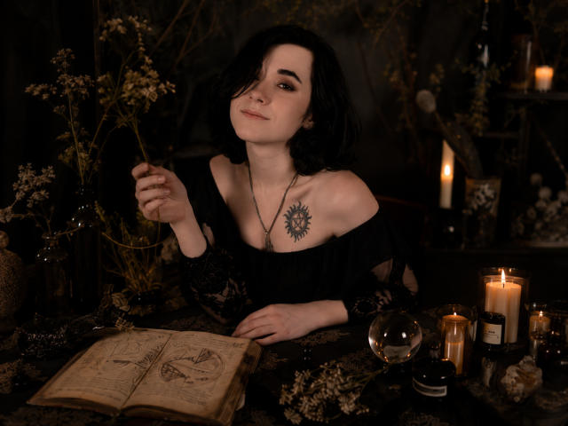 ReginaMay mit dunklem Haar, schwarzem Spitzenoberteil mit Schulterauschnitt, Sonnen-Tattoo auf der Brust, hält eine Blume, Kerzen, ein Buch, eine Glaskugel und eine schwarze Flasche auf einem dunklen, mystischen Tisch..