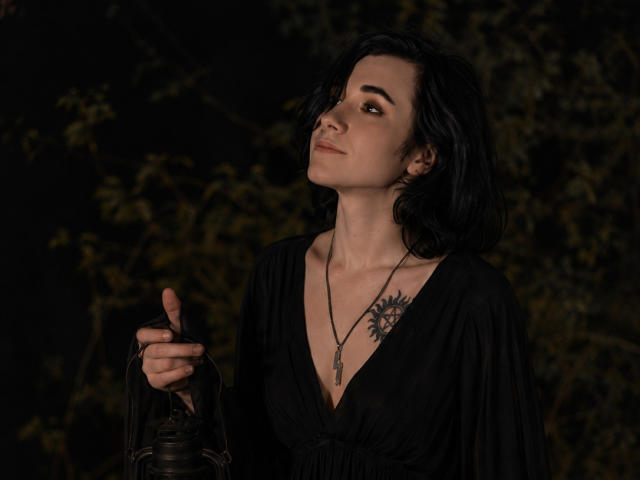 ReginaMay, con cabello negro, mirando hacia arriba, vestida con un vestido negro de escote pronunciado, sosteniendo un farol, mostrando un tatuaje en su pecho, fondo oscuro, noche, sonrisa sutil, collar..
