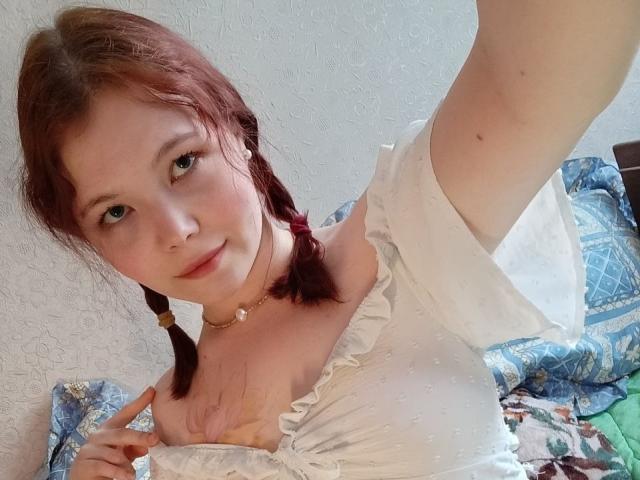 DaynaTays com tranças vermelhas, blusa branca com um ombro à mostra, mostrando os seios com a mão direita, sorrindo, pele clara, olhos verdes, colar e maquiagem leve, em um selfie..
