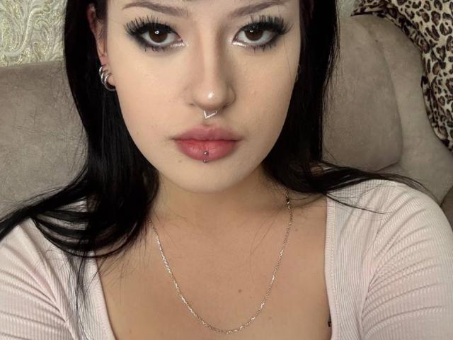 Karrie Pilley hat dunkles Haar, schwarzen Eyeliner, braune Augen, einen Nasenring und ein Lierepiercing mit einer kleinen Kugel. Sie trägt eine silberne Kette und ein pinkes Oberteil..