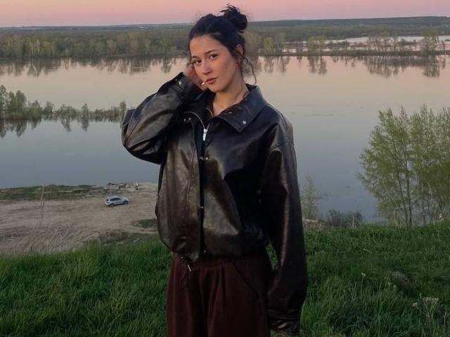 AngleZiak mit dunklem Haar, zu einem unordentlichen Dutt gebunden, trägt eine schwarze Lederjacke und marineblaue Hosen und steht am Ufer eines spiegelnden Sees bei Sonnenuntergang und berührt ihr Haar mit einer Zigarette..