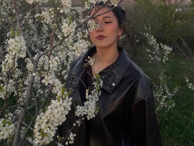 AngleZiak, con i capelli scuri raccolti in uno chignon, indossa una giacca di pelle nera, in piedi di fronte a fiori bianchi, con gli occhi socchiusi, che appare fresca e sicura di sé, con le labbra leggermente tumefatte..