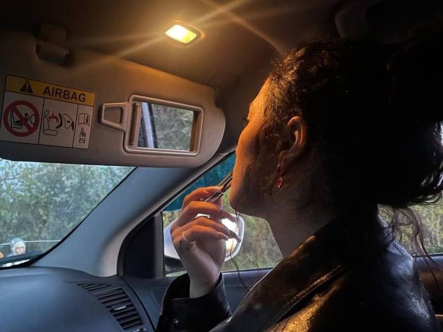 AngleZiak, avec ses cheveux foncés attachés en chignon, portant des boucles d'oreilles rouges et une veste en cuir, sirote une boisson dans un verre transparent à l'intérieur d'une voiture, tenant une cuillère dans sa main droite..