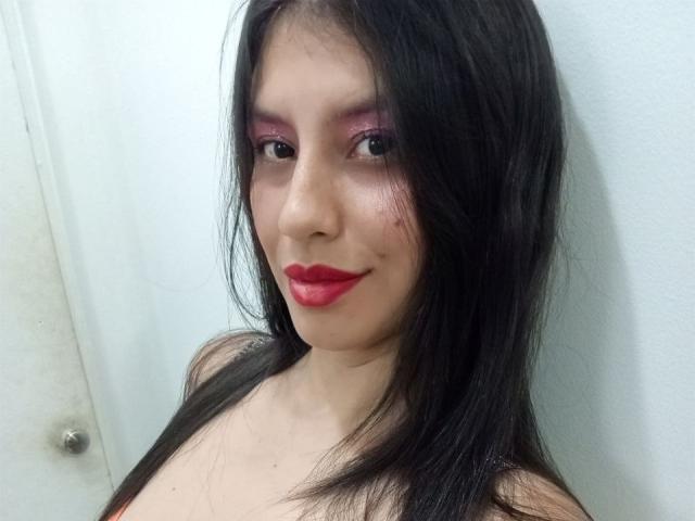 MadissonFontaine has dark straight hair, pink eyeshadow, red lipstick, and a slight smile, showing off her bare shoulders with a subtle smirk. Her skin is light.