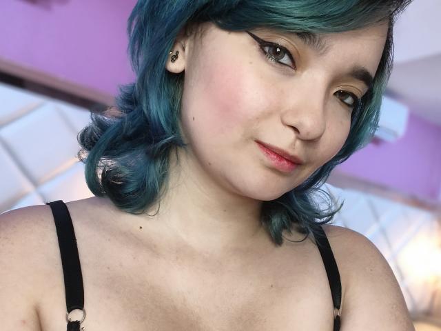 NaommyCarter mit türkisfarbenem Haar, schwarzen Trägern, heller Haut, rosa Rouge, Eyeliner im "Flügel"-Stil, kleiner Nase, rosa Lippen, leichtem Lächeln, kleinen Ohrringen, weichem Licht, entspannter Mimik, leicht sichtbare Oberbrust..