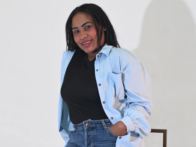 KendalParys ha la pelle scura, lunghi capelli neri, rossetto rosso, indossa una camicia azzurra sopra una maglietta nera e jeans blu, con una mano in tasca, sorridendo alla fotocamera..