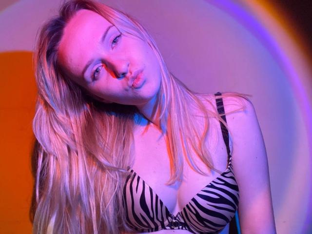 FlyFreedom has blonde hair, pouty lips, and zebra-print tits, looking down with a slight tilt. Pink and blue light casts a sexy glow on her face and chest.