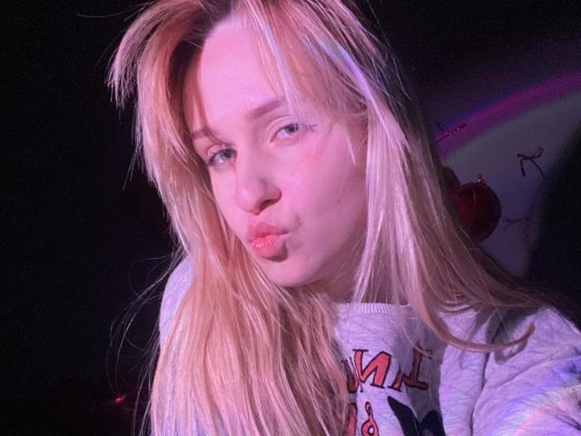 FlyFreedom with pouty pink lips, blonde hair, and a grey hoodie, making a silly face, showing off her cute nose and soft, fair skin. Her tits are not visible.