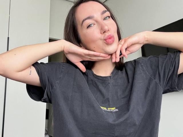 AraiaSheri with puffy lips, black tee, and hands under chin, showing a small tattoo on her right arm, and dark brown hair, in a selfie.