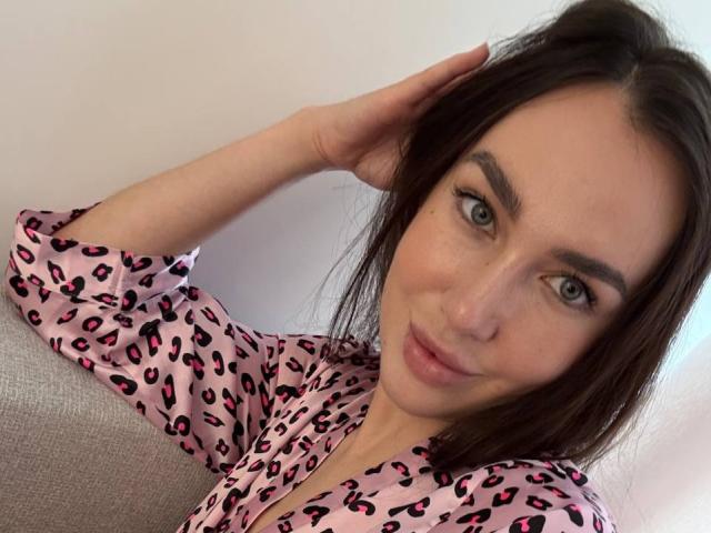 AraiaSheri, con gli occhi azzurri, i capelli castano scuro e una camicia rosa con stampa leopardata, è seduta su un divano grigio, con la testa appoggiata e un leggero sorriso, mentre la sua mano destra tocca i capelli..