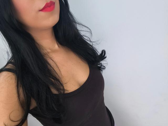 LunaFetish - aantrekkelijk met zwart haar, rode lippenstift en een zwart tanktopje dat haar borsten laat zien, gefotografeerd vanuit de zijkant. Haar huid is licht en ze kijkt iets naar beneden..