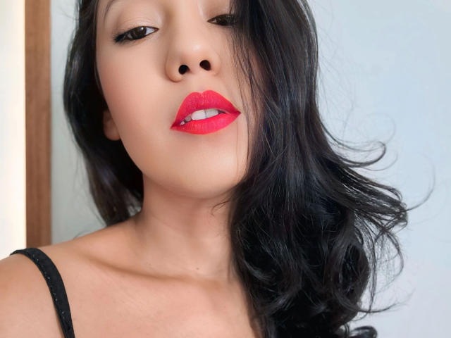 LunaFetish - sensual, com cabelo preto brilhante, batom vermelho vibrante e delineador preto, olhando para cima com os lábios vermelhos levemente entreabertos. Ela está usando uma alça preta. Pele clara e suave..