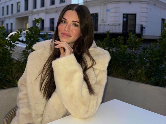 CherryMuse – sinnlich, mit langen braunen Haaren, trägt einen flauschigen weißen Mantel, sitzt an einem weißen Tisch, die Hand nahe am Mund, ein leichtes Lächeln, grüne Augen und natürliches Make-up..