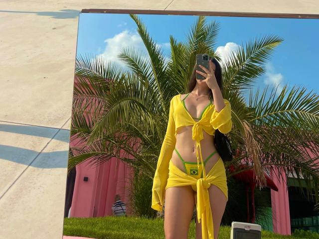 CherryMuse, en maillot de bain jaune composé d'un haut et d'une jupe, dévoile sa lingerie verte à bretelles et un petit tatouage représentant le drapeau brésilien, et prend un selfie devant un miroir avec son téléphone..