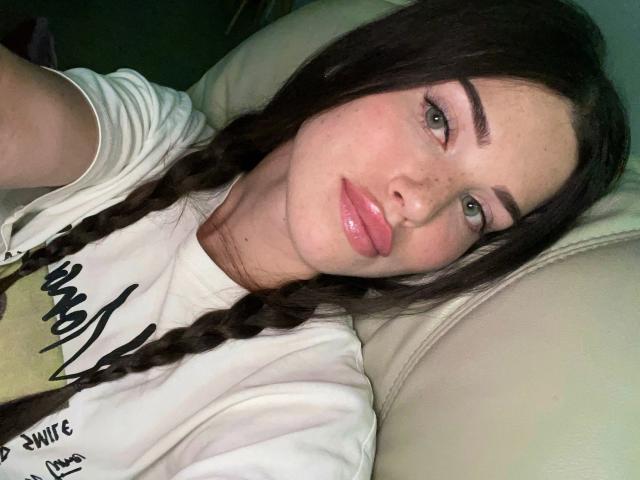CherryMuse, com lábios rosados e volumosos, olhos verdes e sardas, tem tranças castanho-escuras, veste uma camisa branca e está deitada em um sofá bege, olhando para a câmera..