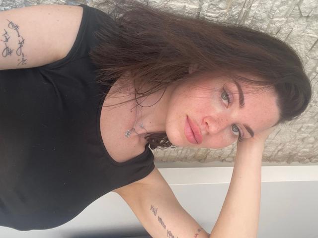 CherryMuse, avec ses cheveux bruns ondulés, son haut noir, ses tatouages sur les bras, ses yeux verts et un léger sourire, reposant la tête sur sa main, affichant une expression détendue et mignonne..