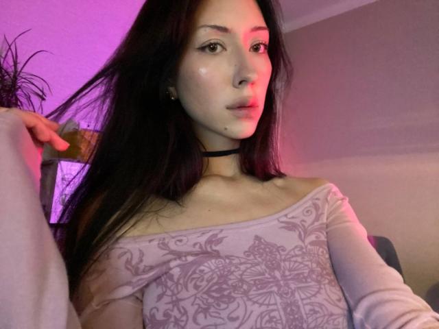Maryln Werntz mit langem, dunklem Haar, trägt ein weißes Oberteil mit Schulterauschnitt und floralen Mustern, hält ihre Haare, im Hintergrund sind lila und rosa Lichteffekte, sie trägt eine schwarze Halskette und dezentes Make-up und blickt in die Kamera..