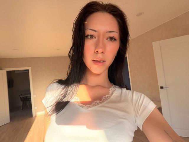 Maryln Werntz, com cabelos pretos longos, pele clara e olhos castanhos, vestindo uma camisa branca com detalhes em renda, iluminada pelo sol em um quarto, mostrando seios pequenos e um sorriso discreto..