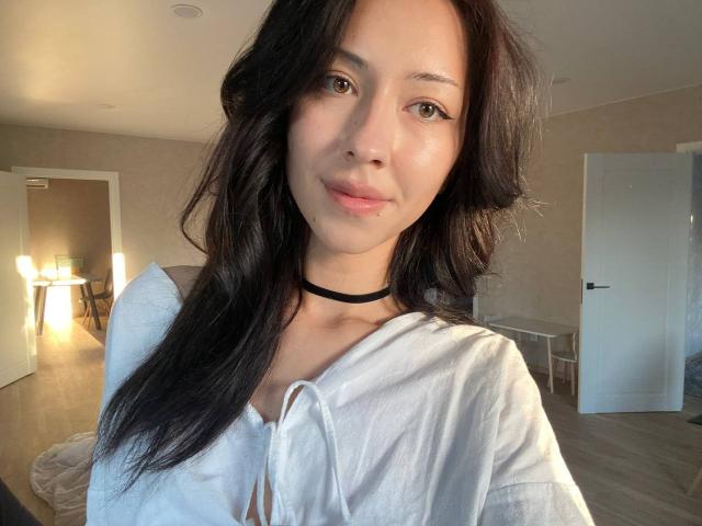 Maryln Werntz, avec de longs cheveux noirs ondulés, portant une chemise blanche et un collier noir, souriant doucement, avec des yeux marrons, une peau claire et de petites seins, dans une pièce baignée de soleil..
